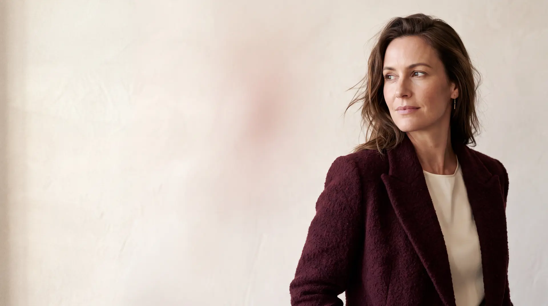 A woman in a burgundy jacket looking thoughtfully to one side against a soft plaster wall